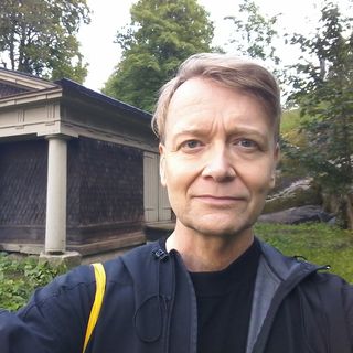 Kuvassa Lasse Majuri katsoo suoraan kameraan. Taustalla näkyy vehreää luontoa ja kaunis rakennus, jossa on näyttävä vaalea pylväs kulmassa.